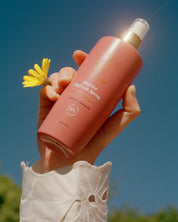 Hand holding a curly angels rejoice refresh spray bottle with a yellow flower against a blue sky