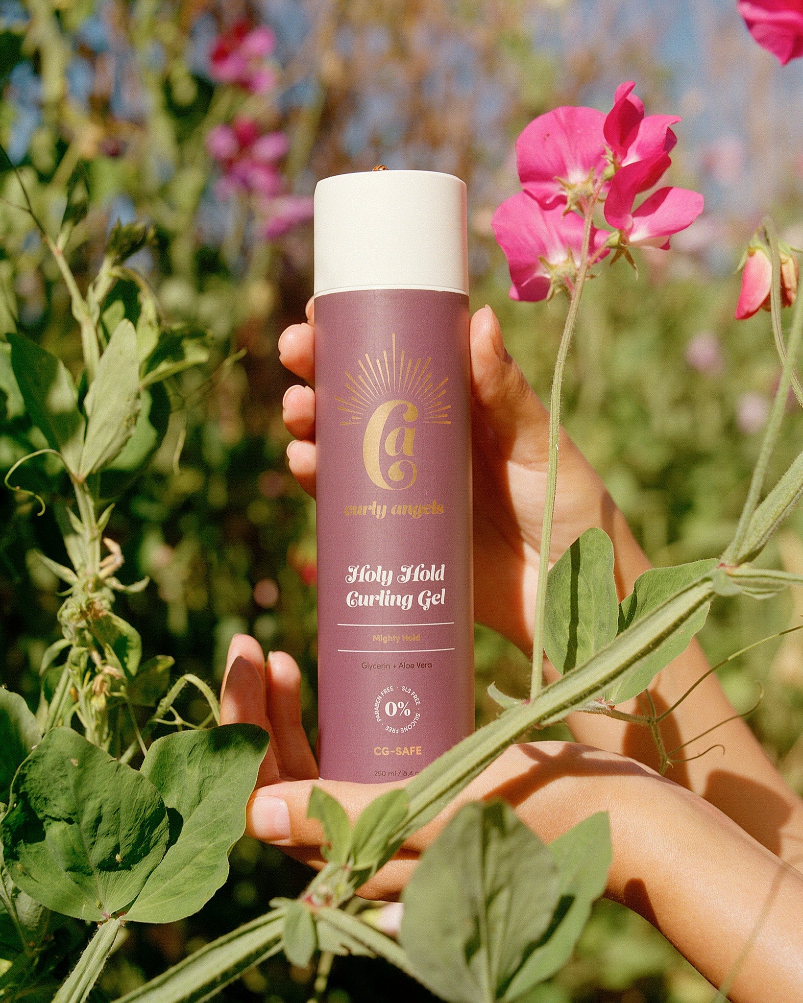 Hand holding a bottle of curly angels Holy Hold curling gel among green leaves and pink sweet pea flowers - vegan - cruelty-free
