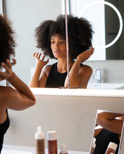 Woman with Afro styling her hair in front of a mirror with Curly Angels Glorious Curling Gel on the counter.