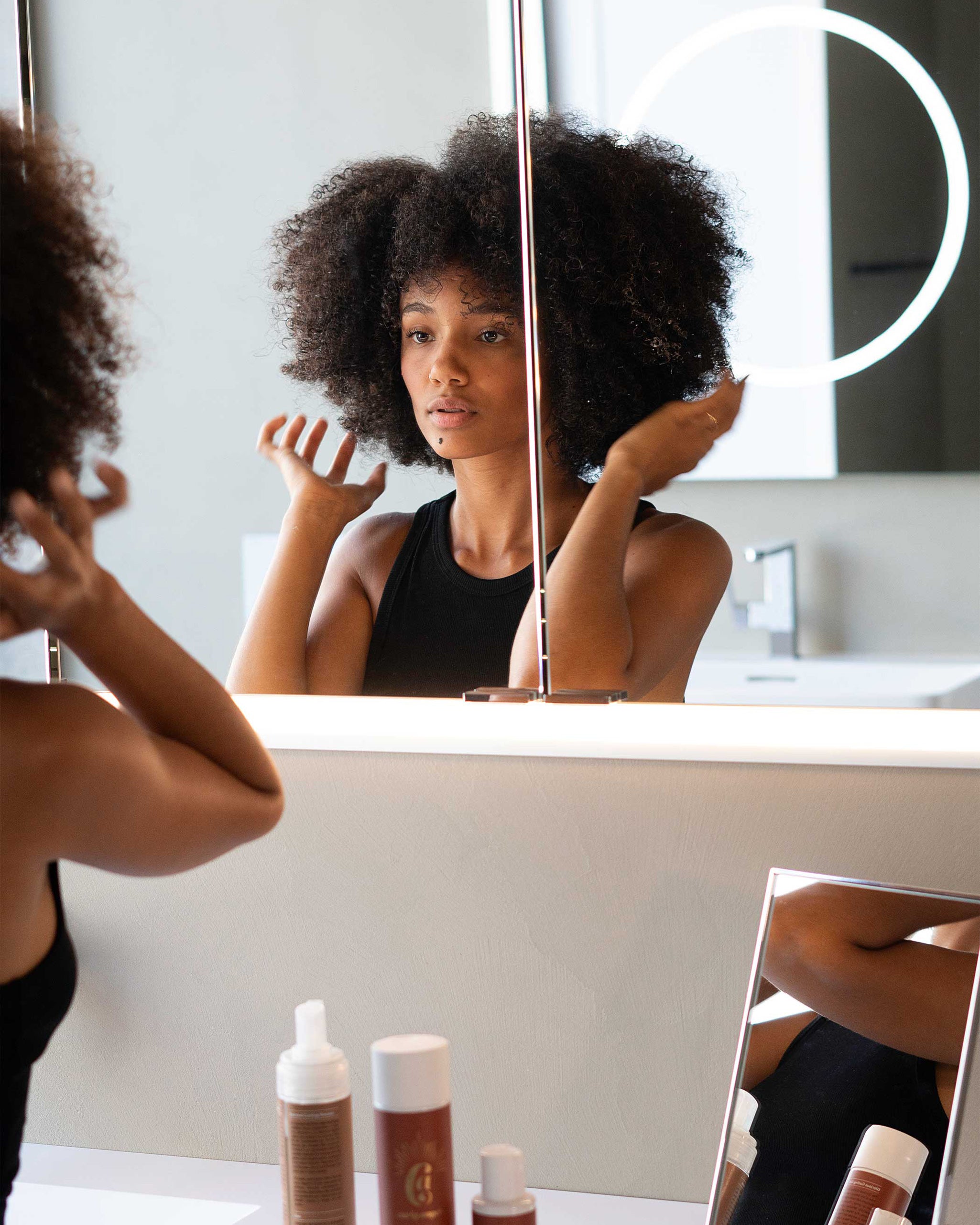 Woman with Afro styling her hair in front of a mirror with Curly Angels Glorious Curling Gel on the counter.