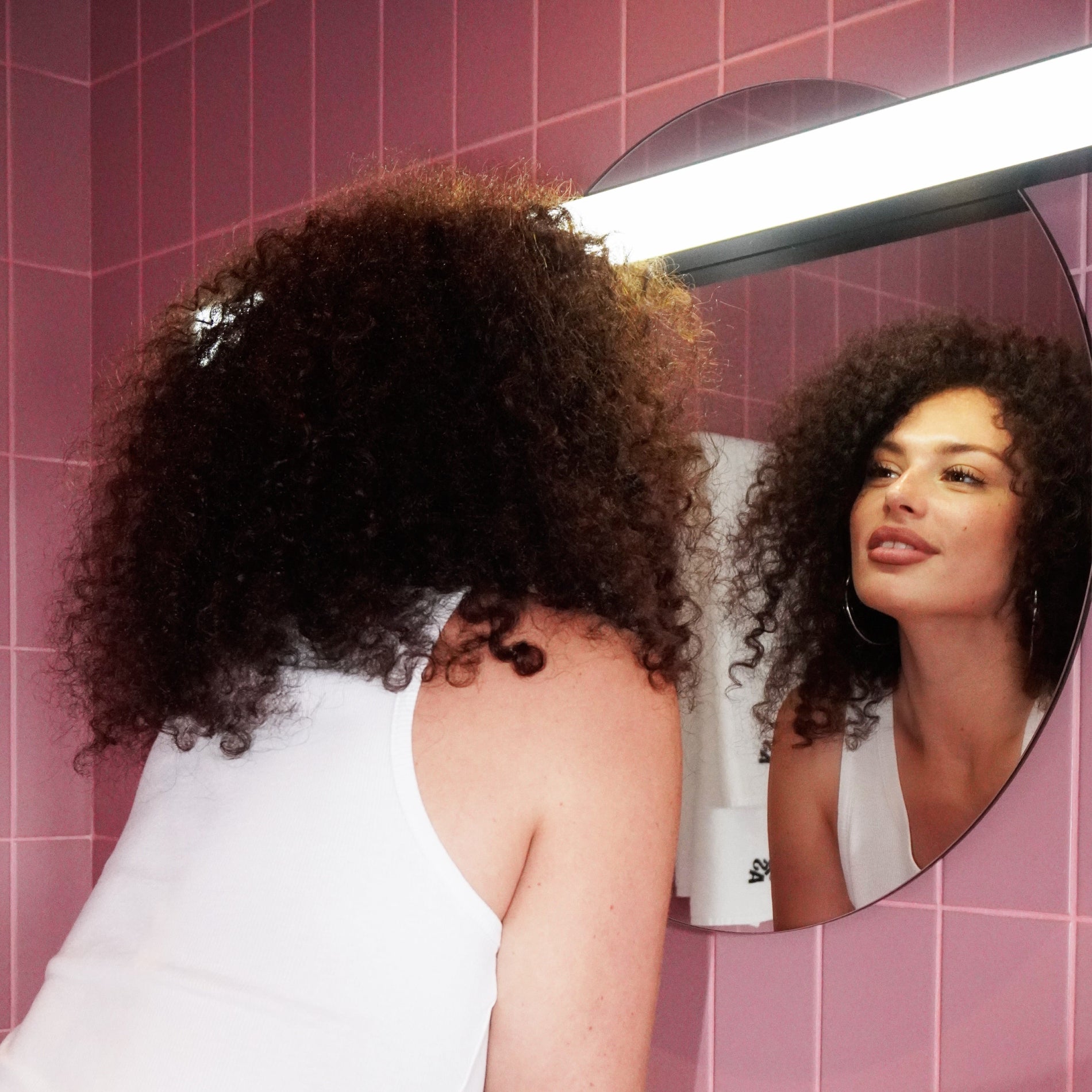 Woman with curly hair looking at herself in a round mirror against a pink tiled wall.