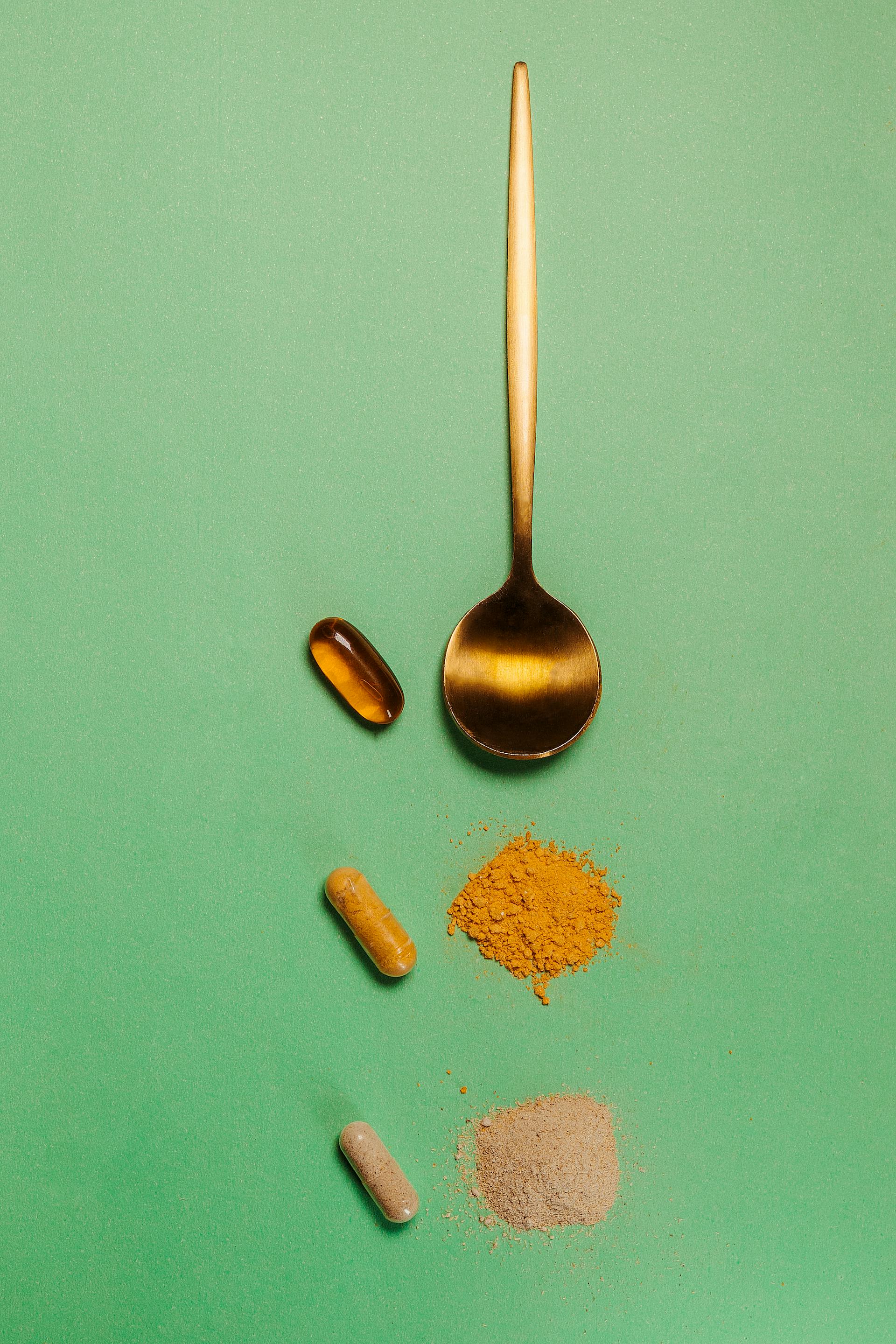 Gold spoon with plant powders, oil pill, and capsules on a green background